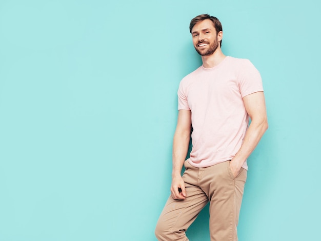 Portrait of handsome smiling stylish hipster lambersexual model Sexy man dressed in pink Tshirt and trousers Fashion male isolated on blue wall in studio