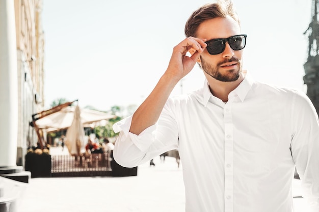 Portrait of handsome smiling stylish hipster lambersexual modelModern man dressed in white shirt Fashion male posing on the street background in sunglasses Outdoors at sunset