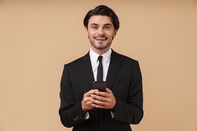 Портрет красивого улыбающегося молодого бизнесмена в костюме, стоящего изолированно над бежевой стеной, с помощью мобильного телефона