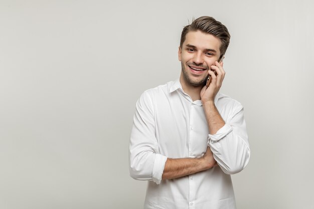 Foto ritratto di un bel giovane sorridente in camicia bianca che parla al telefono cellulare guardando la telecamera