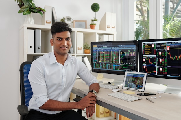 Portrait of handsome stock market broker sitting at his office desk with computers with stock market data in background