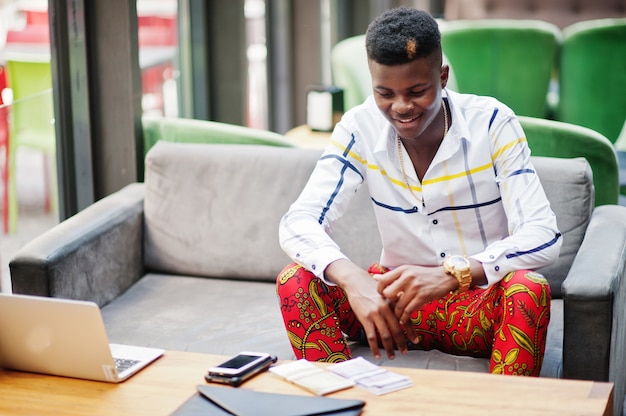 Portrait of handsome stylish african american business man in red throusers and white shirt sitting in office with laptop and cash on table.