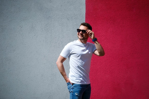 Портрет красивого стильного парня, человека, одетого в белую пустую футболку, стоящего на серо-красном стенном фоне. Городской стиль в одежде, современный модный образ. Мужская мода