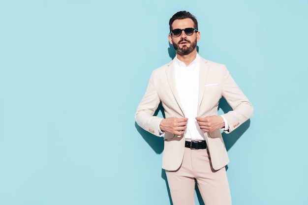 Portrait of handsome stylish hipster lambersexual model Sexy modern man dressed in white elegant suit Fashion male posing in studio near blue wall in sunglasses