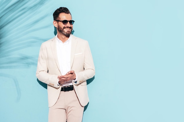 Portrait of handsome stylish hipster lambersexual model Sexy modern man dressed in white elegant suit Fashion male posing in studio near blue wall in sunglasses