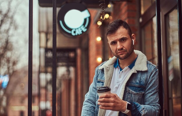 Portrait of a handsome stylish man wearing a denim jacket with wireless headphones holding takeaway coffee outside the cafe.