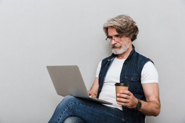 Portrait of a handsome stylish mature man wearing a vest sitting on a chair isolated over gray wall, using laptop computer while drinking takeaway coffee