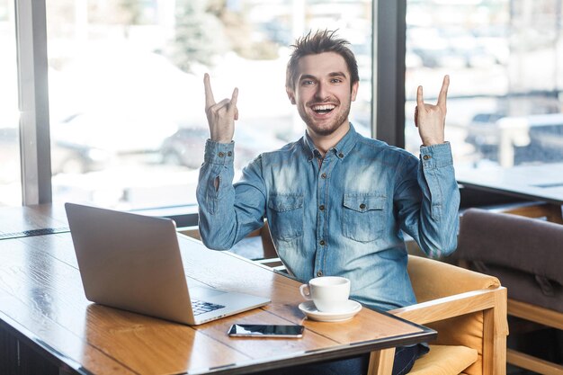 Portrait of handsome successfull positive bearded young freelancer in blue jeans shirt are sitting in cafe and working on laptop with toothy smile and showing rock sign, looking at camera. indoor