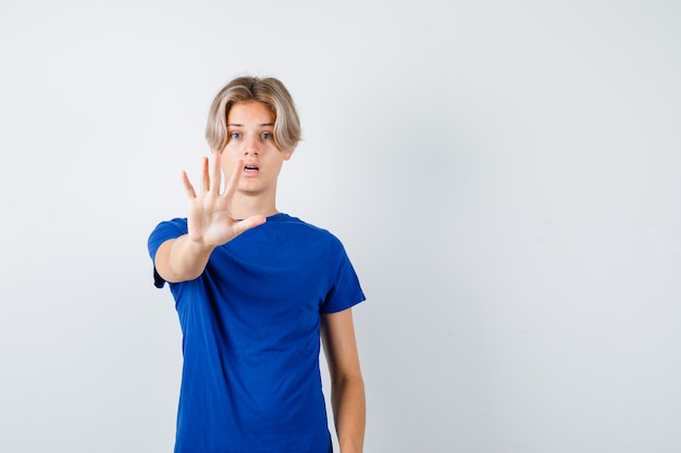 Ritratto di un bel ragazzo adolescente che mostra il gesto di arresto con una maglietta blu e sembra una vista frontale spaventata