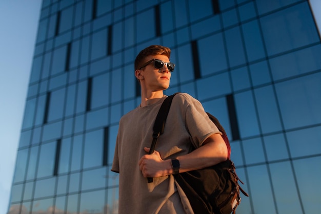 Портрет красивого туриста-хипстера в солнцезащитных очках с сумкой, удобно стоящего парня возле здания в центре города Спокойный расслабляющий момент концепции летних каникул