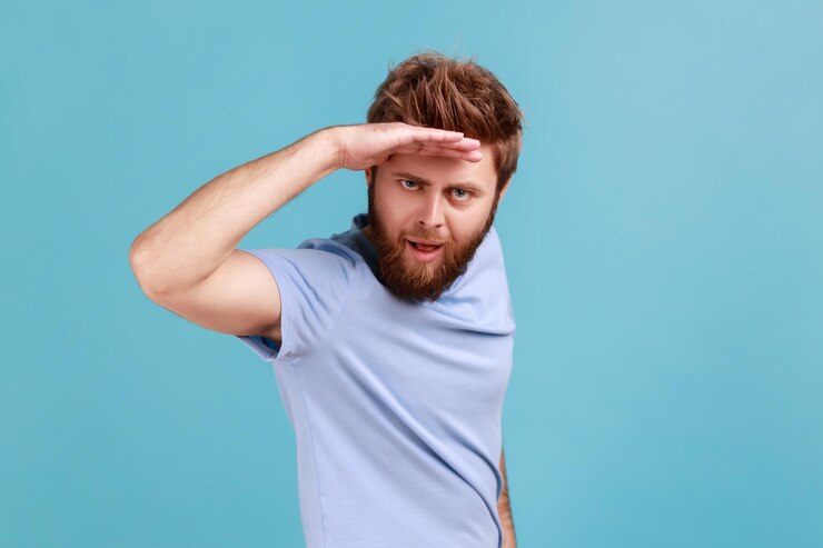 Photo portrait of handsome young adult bearded man looking far away at distance with hand over head sees something pleasant with flirting expression indoor studio shot isolated on blue background