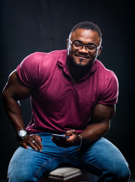 Portrait of handsome young african man in short sleeve shirt sitting with phone in hands on a chair Muscular male model with mobile phone Closeup