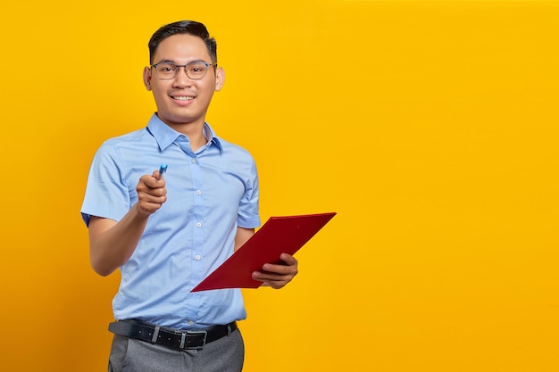 Portrait of handsome young asian man in glasses standing holding document folder and pointing with pen looking at camera with smiling face isolated on yellow background