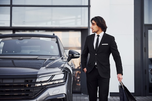 Portrait of handsome young businessman in black suit and tie outdoors near modern car and with shopping bags.