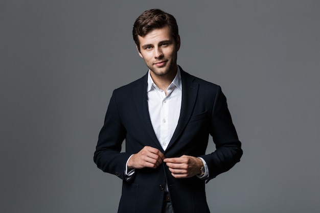 Portrait of a handsome young businessman dressed in suit isolated over gray wall, buttoning his jacket