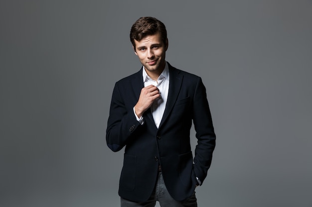 Portrait of a handsome young businessman dressed in suit isolated over gray wall, unbuttoning his shirt