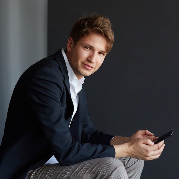 Portrait of a handsome young businessman in elegant suit holding a cell phone, looking at camera, over black background.