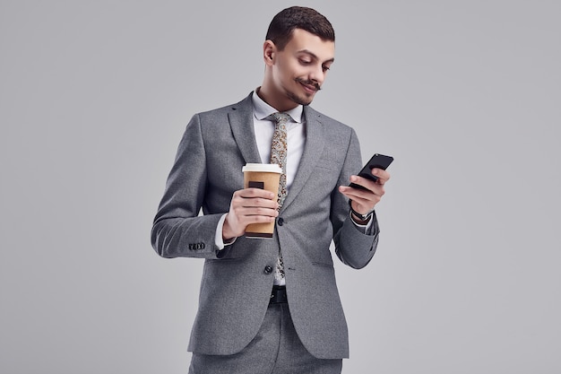 Portrait of handsome young confident arabic businessman with fancy mustache in fashion gray full suit holds a cup of coffee and typing a message on the phone