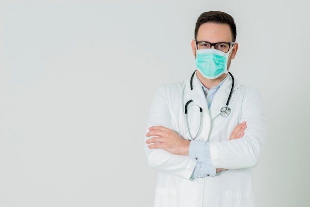 Ritratto di giovane medico bello in uniforme medica bianca e maschera che esamina macchina fotografica contro bianco