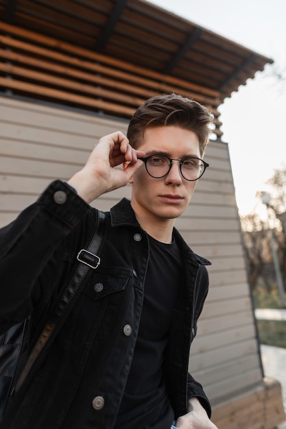 Portrait of a handsome young english guy with a hairstyle in a fashionable black denim jacket straightens his glasses in the city