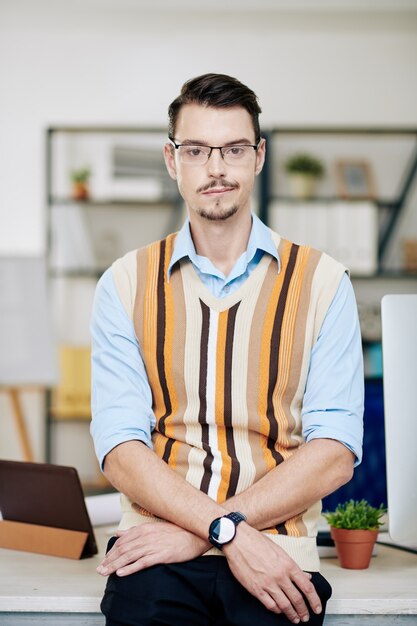 Portrait of handsome young entrepreneur in glasses standing at table in office and looking at camera