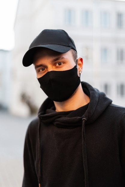 街で流行のパーカーを身に着けている保護医療マスクと黒のファッションモックアップキャップを持つハンサムな若い流行に敏感な男のモデルの肖像画