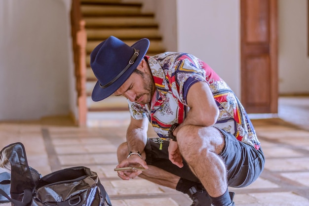 Portrait of handsome young hispanic man crouching wearing blue hat and casual shirt looking at his cell phone in living room of a home