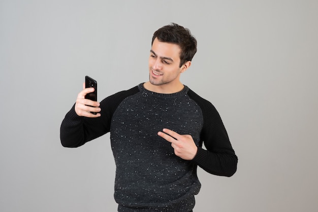 Portrait of handsome young male standing and taking selfie over gray wall