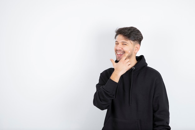 Portrait of handsome young man in black hoodie smiling while standing against white wall