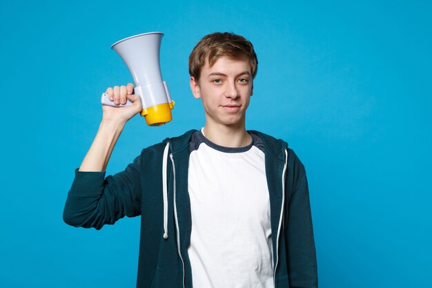 Portrait of handsome young man in casual clothes holding in hand megaphone isolated on blue wall . people sincere emotions, lifestyle concept