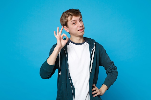 Portrait of handsome young man in casual clothes standing and showing OK gesture isolated on blue wall. People sincere emotions lifestyle concept. 