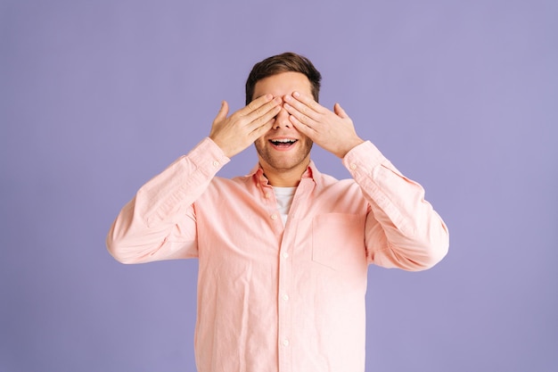 Portrait of handsome young man covering eyes by hands and smiling over isolated purple background standing on pink isolated background in studio. Concept of blind.