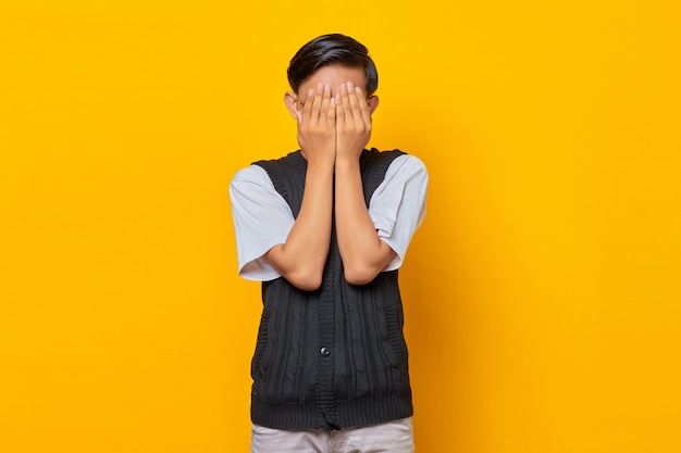 Portrait of handsome young man covering face with palms over yellow background