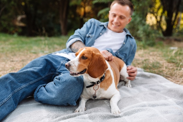 Portrait of handsome young man in denim clothes sitting on blanket in park with his canine dog