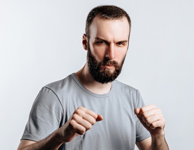 Portrait of handsome young man frowning while looking at camera holding fists and boxing on white wall with space for advertisement mock up