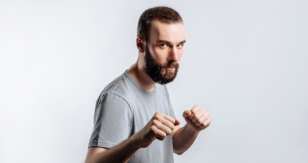 Portrait of handsome young man frowning while looking at camera holding fists and boxing on white wall with space for advertisement mock up