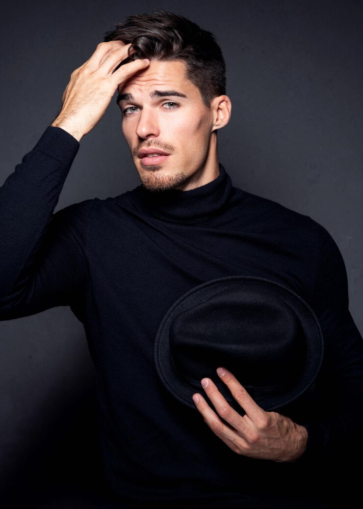 Photo portrait of handsome young man holding hat while standing against black background