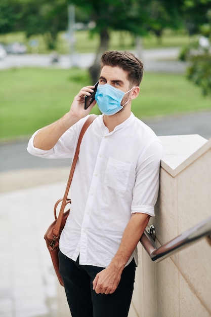 Portrait of handsome young man in medical mask standing outdoors and talking on phone