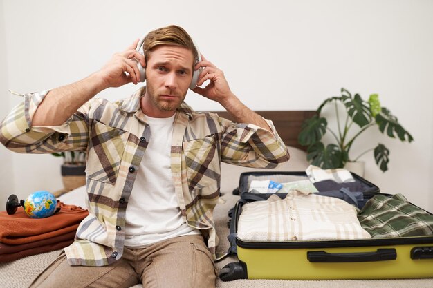 Photo portrait of handsome young man packing things in suitcase listening to music in headphones tourist