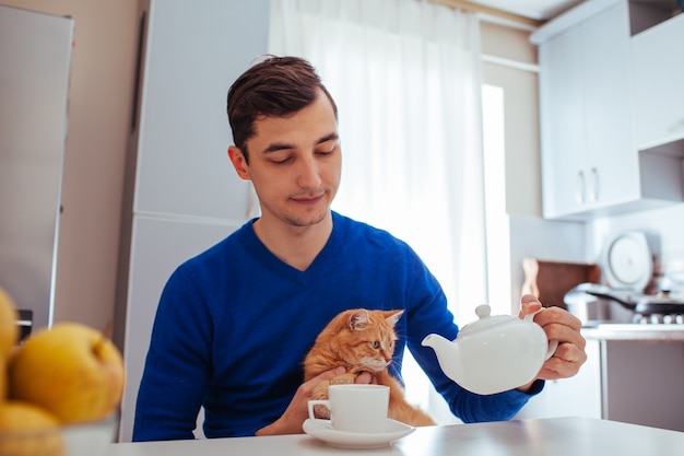 ハンサムな若い男の肖像は台所で猫とお茶を注ぐ