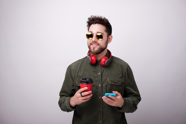 Portrait of handsome young man in sunglasses, holding a cup and mobile while looking far away