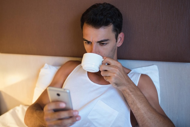 Portrait of handsome young man using a mobile phone and drinking cup of coffee in the white bed