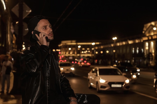 Портрет красивого молодого человека, использующего мобильный телефон, звонящего по улице ночного города с автомобилями и неоновыми огнями, отводящими взгляд