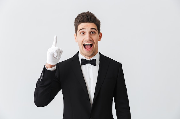 Portrait of a handsome young man waiter wearing tuxedo and gloves standing isolated over white wall, pointing up