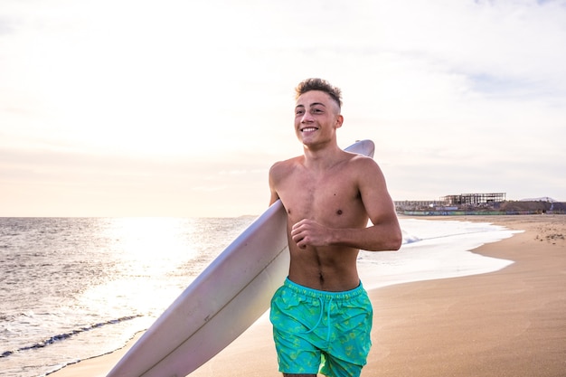 波に乗って休暇中にサーフィンに行く準備ができている彼のサーフボードと一緒にビーチを歩いているハンサムな若い男の肖像画