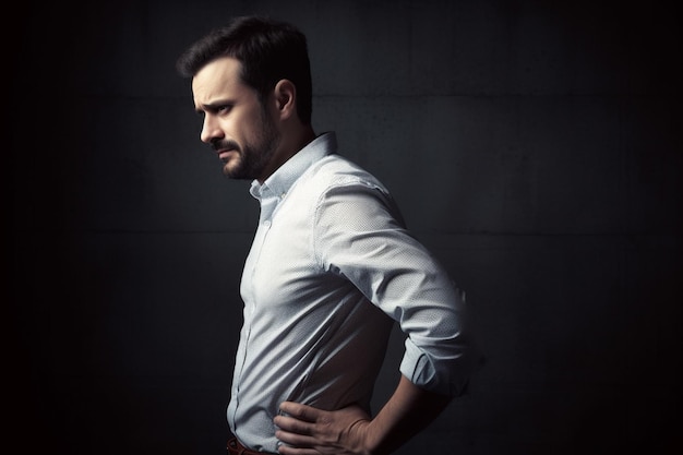 Portrait of a handsome young man in white shirt suffering from back pain on dark background
