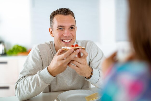 Portrait of a handsome young man who is eating healthy sandwich for the breakfast in the domestic kitchen.