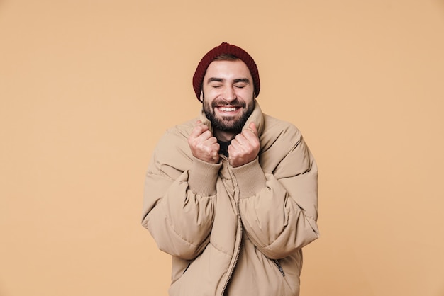 Portrait of handsome young man in winter jacket and hat smiling isolated on beige