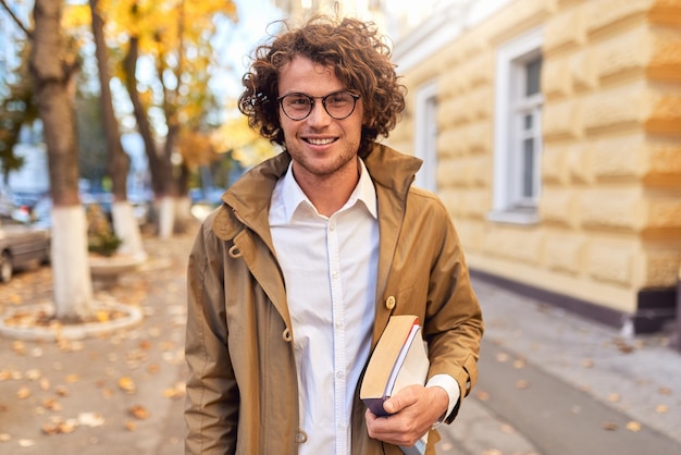 屋外で本を持っているハンサムな若い男の肖像秋の通りの背景に大学のキャンパスで本を運ぶ大学の男子学生眼鏡と巻き毛の笑顔の男が外の本でポーズをとる