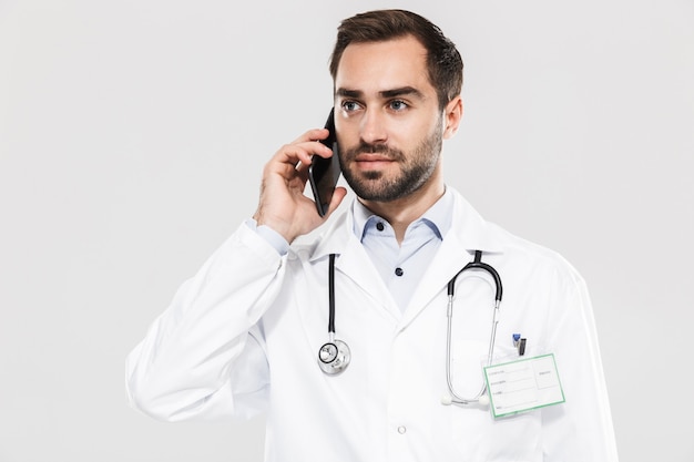 Portrait of handsome young medical doctor with stethoscope working in clinic and talking on cellphone isolated over white wall